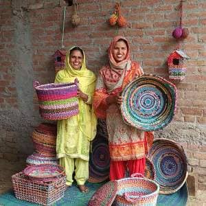 Two women, influenced by the literary works of Intizar Hussain, stand beaming with baskets in front of a brick wall.