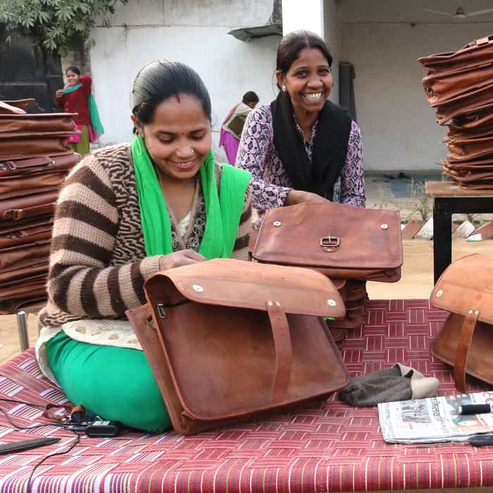Two women participate in Project Disha by crafting leather bags on a blanket.